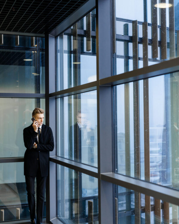 Young man standing by window of office-center and talking by cellphone